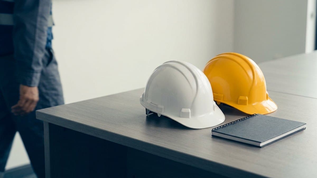 A white and a yellow hard hat resting on a desk beside a closed notebook